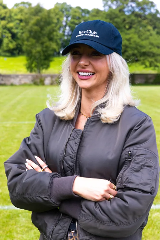 Rex Club | A smiling blonde woman wears a navy Rex Club Sports Headwear Dad Cap and a dark bomber jacket, standing outdoors on grass with trees and a stone wall in the background. | Custom Caps | Custom Hats | Team Headwear | UK