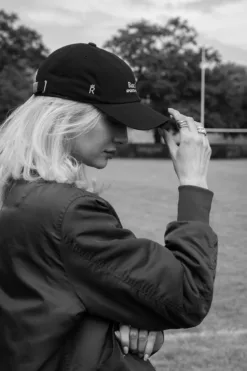 Rex Club | Black and white photo of a woman outdoors, wearing the Rex Club Sports Headwear Dad Cap and a jacket, adjusting her cap with one hand. Trees and a grass field appear in the background. | Custom Caps | Custom Hats | Team Headwear | UK
