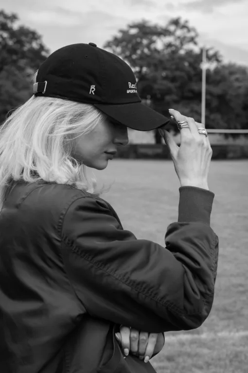 Rex Club | Black and white photo of a woman outdoors, wearing the Rex Club Sports Headwear Dad Cap and a jacket, adjusting her cap with one hand. Trees and a grass field appear in the background. | Custom Caps | Custom Hats | Team Headwear | UK