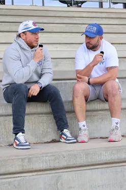 Rex Club | Two men sit on outdoor steps, chatting with microphones. One wears a gray hoodie and cap; the other wears a white shirt, shorts, and a blue Rex Club Sports Headwear Trucker cap. | Custom Caps | Custom Hats | Team Headwear | UK