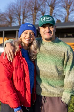 Rex Club | A smiling man in a green hat and sweater stands outdoors with his arm around a woman wearing a red jacket and the Rex Club Sports Headwear Beanie in blue, posing together on a sunny day with trees and a building behind them. | Custom Caps | Custom Hats | Team Headwear | UK