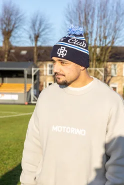Rex Club | A man wearing a Rex Club Bobble Hat and a light-colored "MOTORINO" sweater stands outdoors on a sunlit grassy field with trees and buildings in the background. | Custom Caps | Custom Hats | Team Headwear | UK