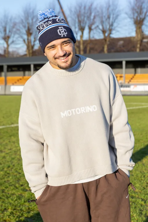 Rex Club | A man wearing a beige "MOTORINO" sweater, brown pants, and the Rex Club Bobble Hat stands smiling on a grassy field with yellow stadium seats and leafless trees in the background. | Custom Caps | Custom Hats | Team Headwear | UK