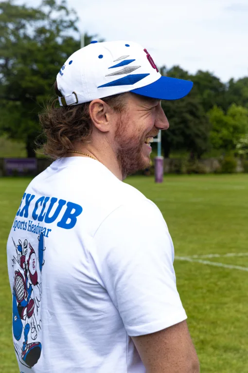 Rex Club | A man in a white Rex Club - Summer 24 - Slouch cap and T-shirt with a sports cartoon and "CLUB" on the back stands smiling on grass, trees behind him. | Custom Caps | Custom Hats | Team Headwear | UK