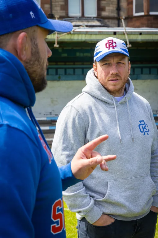 Rex Club | Two men in casual sportswear chat outside a building. One, dressed in the Rex Club - Summer 24 - Slouch gray hoodie and white cap, listens with hands in pockets as his friend gestures while speaking. | Custom Caps | Custom Hats | Team Headwear | UK