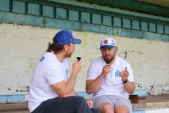 Rex Club | Two men in Rex Club - Summer 24 - Slouch baseball caps and white shirts sit on concrete bleachers, chatting with microphones. Behind them is a weathered blue wall and a metal roof over the seating area. | Custom Caps | Custom Hats | Team Headwear | UK