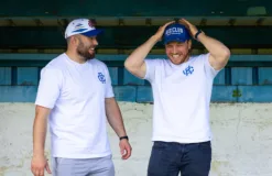 Rex Club | Two men in Rex Club Summer T Shirt 24s and baseball caps smile and laugh together under a blue-green shelter. One adjusts his cap while the other stands with his hands at his sides. | Custom Caps | Custom Hats | Team Headwear | UK