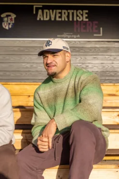 Rex Club | A man in a green sweater and a Rex Club TwoTone Canvas Cap sits on wooden bleachers in sunlight, smiling slightly. Behind him, a sign on a dark wall reads 