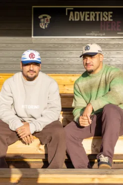 Rex Club | Two men in casual clothes wear Rex Club - TwoTone - Canvas Caps as they sit on wooden benches in sunlight. A sign reading 