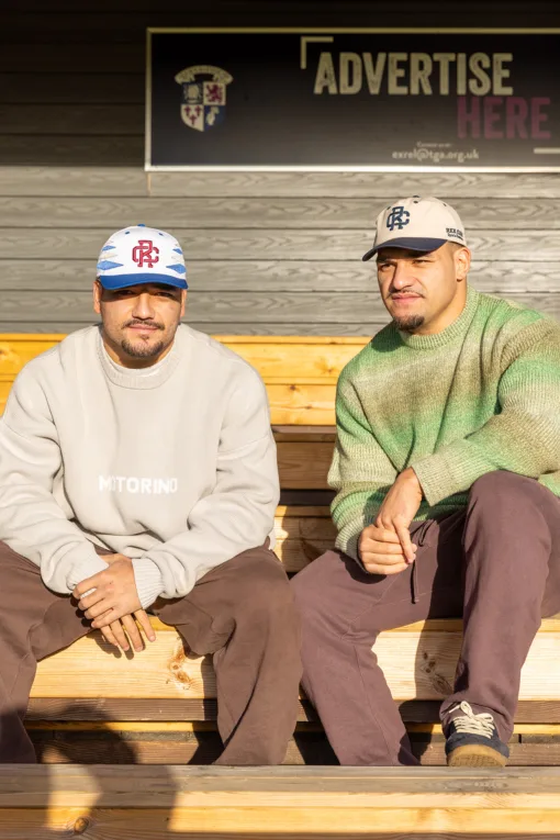 Rex Club | Two men in casual clothes wear Rex Club - TwoTone - Canvas Caps as they sit on wooden benches in sunlight. A sign reading "ADVERTISE HERE" is behind them on a dark wooden wall, and both face the camera. | Custom Caps | Custom Hats | Team Headwear | UK