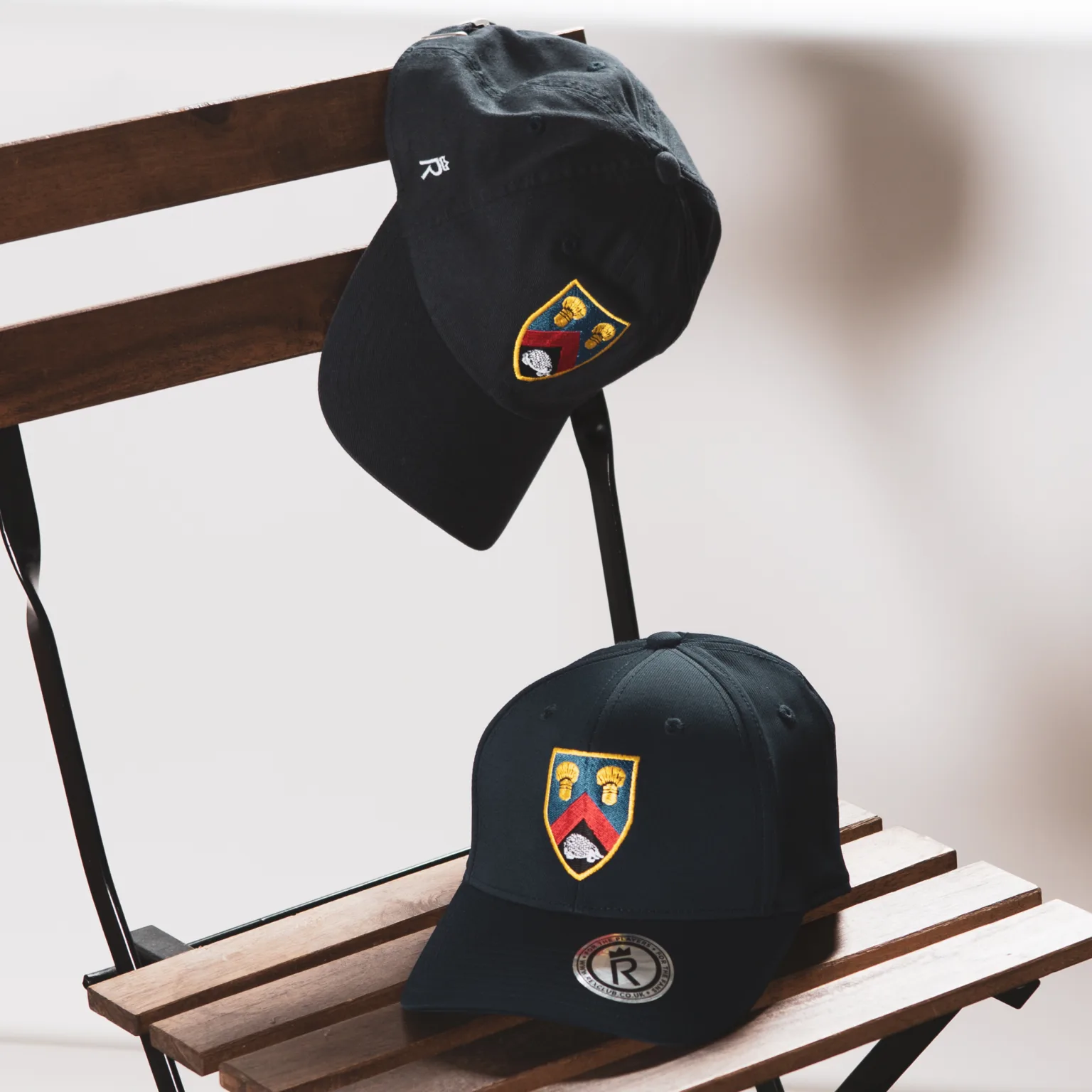 Rex Club | Two black baseball caps with colorful embroidered crests rest on a wooden folding chair; one cap is placed on the seat, and the other hangs over the backrest. | Custom Caps | Custom Hats | Team Headwear | UK
