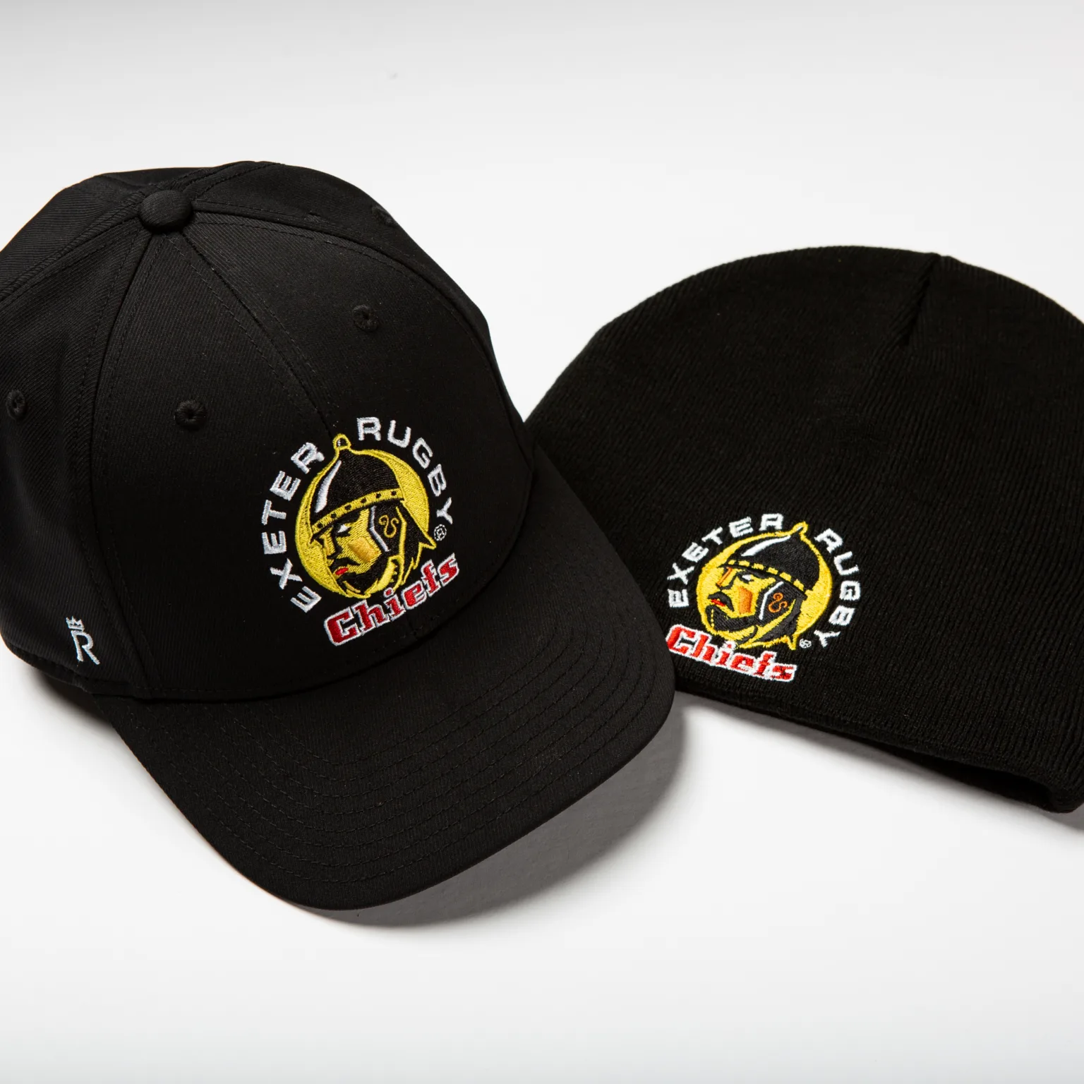 Rex Club | Two black hats—a baseball cap and a knit beanie—both featuring the Exeter Rugby Chiefs logo with a yellow and red chief's head and text embroidered on the front, placed on a white surface. | Custom Caps | Custom Hats | Team Headwear | UK