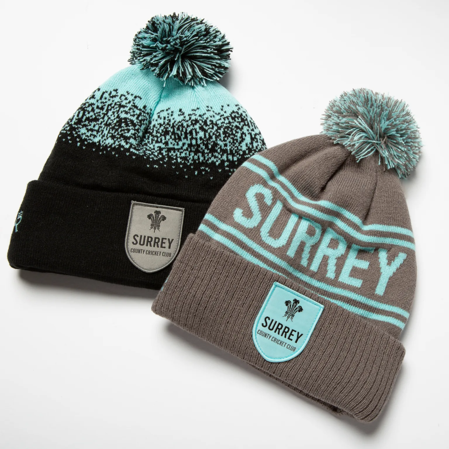 Rex Club | Two knit beanies with pom-poms, both featuring "Surrey County Cricket Club" logos. One is black with turquoise details; the other is gray with turquoise and white stripes. Both have a club crest patch on the front. | Custom Caps | Custom Hats | Team Headwear | UK