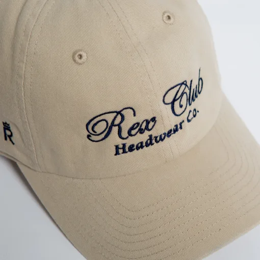 Rex Club | A close-up of a beige baseball cap with "Rex Club Headwear Co." embroidered in navy blue script on the front. | Custom Caps | Custom Hats | Team Headwear | UK
