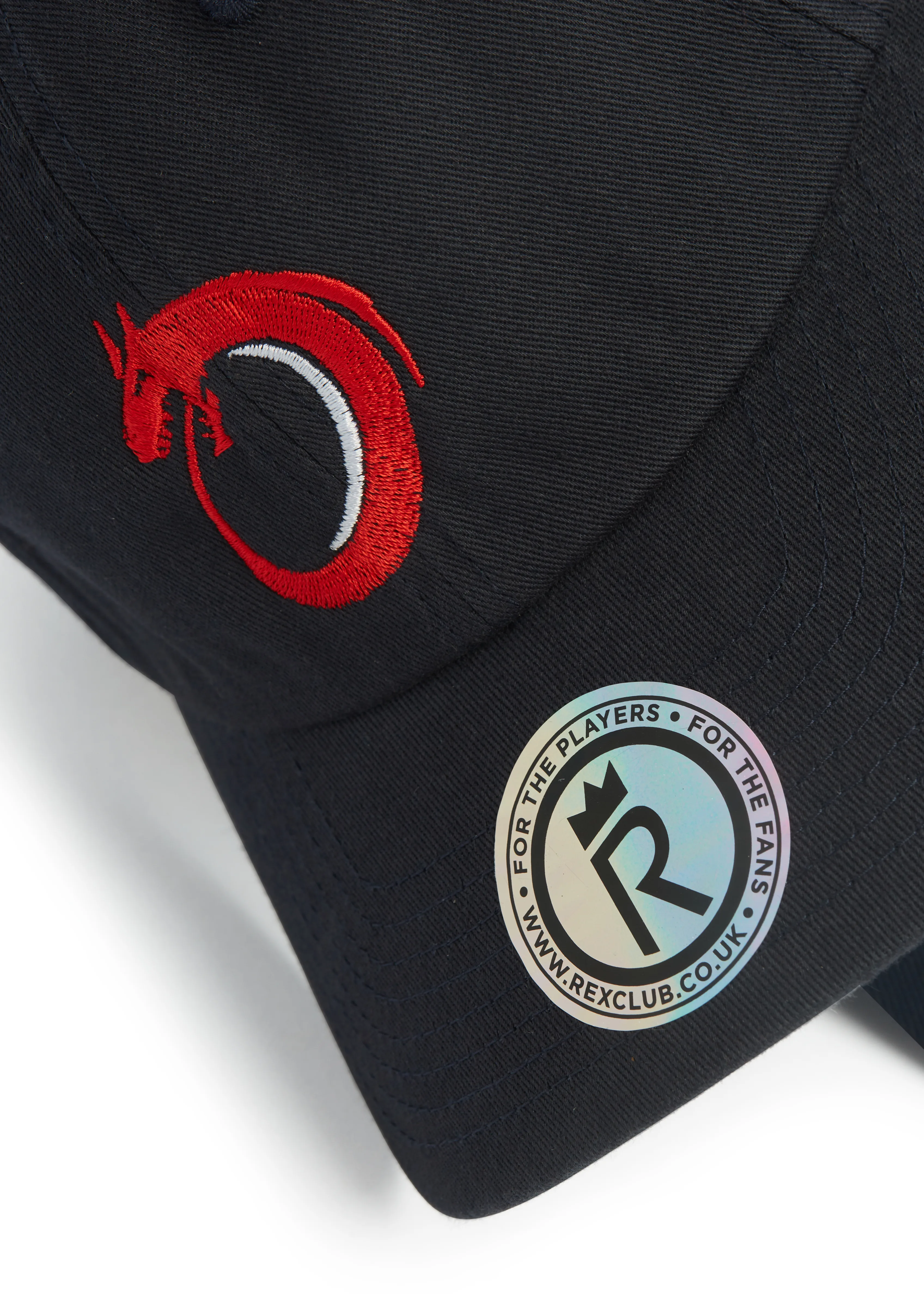 Rex Club | Norway Rugby Slouch | Custom Caps | Custom Hats | Team Headwear | UK Rex Club | A close-up of the Norway Rugby Stretch black cap with a red dragon embroidered logo and a round sticker reading “FOR THE PLAYERS • FOR THE FANS” and “WWW.REXCLUB.CO.UK” around a stylized "R" and crown. | Custom Caps | Custom Hats | Team Headwear | UK