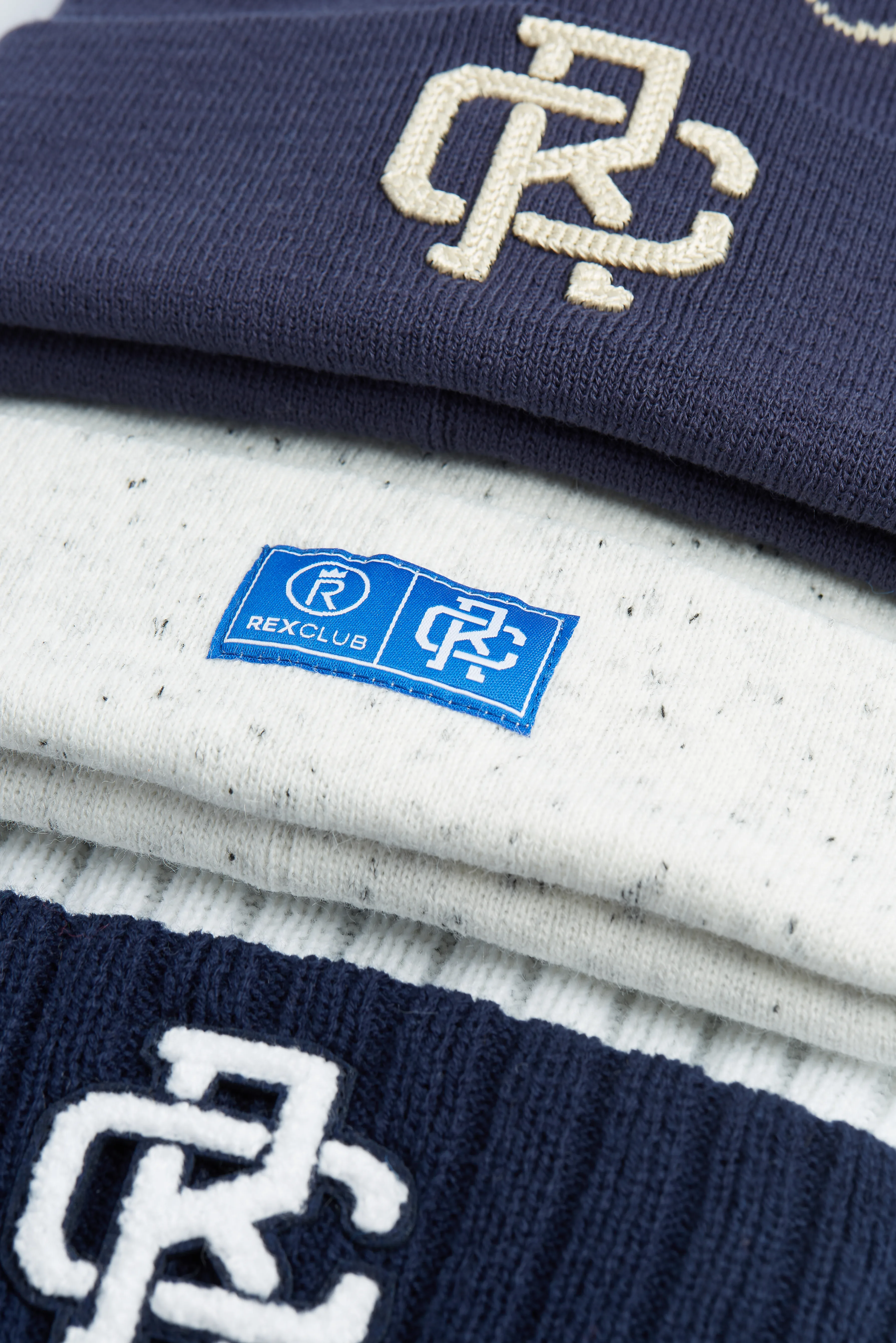 Rex Club | A close-up of folded knitwear, including navy and white pieces. The navy knits have a white "RC" logo, and the white knit has a blue "REXCLUB" label with similar branding. | Custom Caps | Custom Hats | Team Headwear | UK