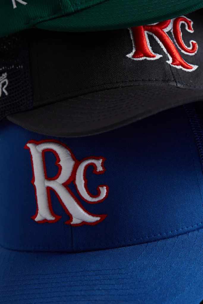 Rex Club | A close-up of three baseball caps stacked on top of each other in green, black, and blue, each featuring a white and red "RC" logo embroidered on the front. | Custom Caps | Custom Hats | Team Headwear | UK