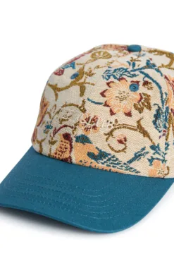 Close-up of a curved-teal brim cap with a floral tapestry-patterned crown on beige fabric.